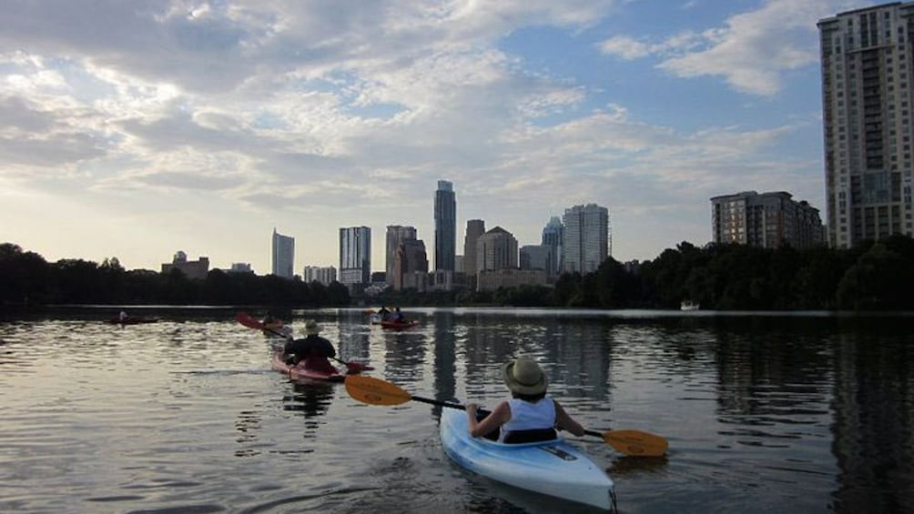 Kayakers shown from behind paddling over river with view of skyline.