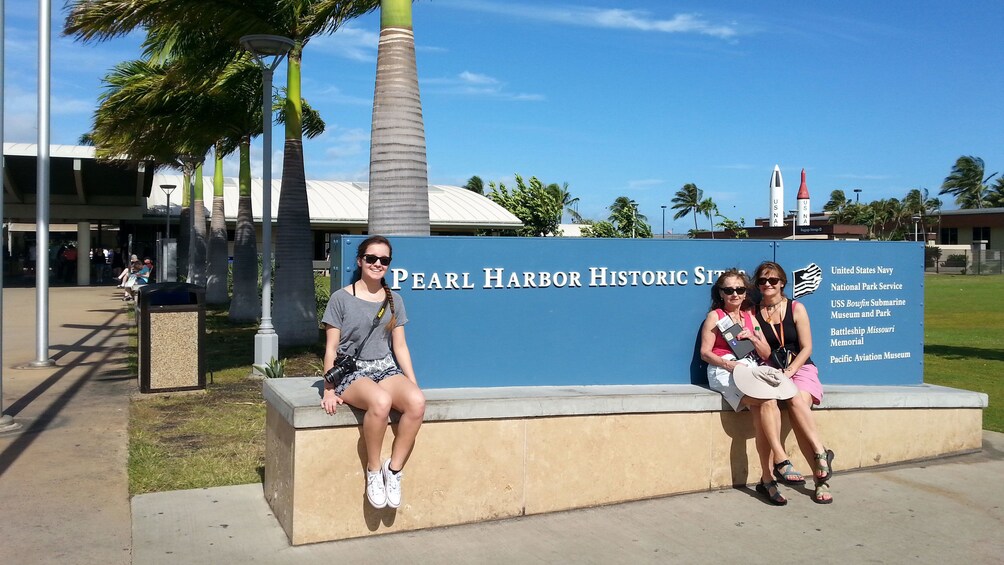 Three women sit next to Pearl Harbor Historic Site sign in Oahu