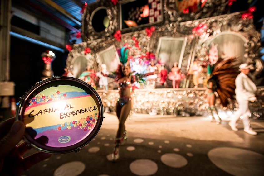 Hand holds small Carnaval Experience emblem with intricate Carnaval exhibit in the background