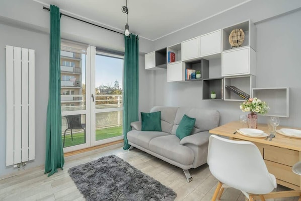 A bright living room with large windows, a gray sofa, green pillows, and a small wooden desk beside it.
