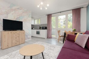 Another perspective of the main room featuring the burgundy sofa, a wooden TV cabinet against a subtle marble-effect wall, and pink curtains leading to the kitchen.
