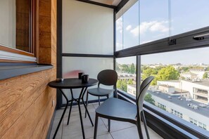 A cozy balcony with a glass railing, featuring a small black table and two chairs, overlooking the city.


