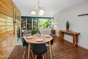 A cozy dining area with a round wooden table set for four surrounded by black and light wooden chairs. The room features a wooden paneled wall, a fireplace, and large windows with a view of greenery.