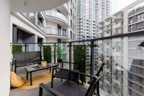 A spacious balcony with outdoor seating, plants, and glass railings, offering a relaxing urban retreat.