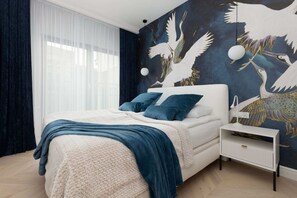 A stylish bedroom with a bold wallpaper design, a blue bedspread, and a large window allowing natural light.

