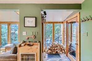 A light-filled room with green walls features a wood rack, a framed picture of a dog, and French doors opening to an outdoor area with a stack of firewood.