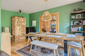 A dining room with green walls features a wooden table with six seats, a wooden cabinet, a bookshelf, and a pendant light. A patterned rug lies under the table, and the room is decorated with various items.