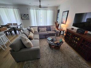 LIVING ROOM: Experience comfort and style in our well-appointed living room, featuring a cozy sofa, a smart TV for entertainment, and a chic center table. Sunlight streams through the large windows perfectly aligned with the adjacent door. It bathes the room in natural light, highlighting the captivating wall paintings in elegant frames.