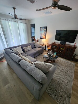 LIVING ROOM: Take in the spacious elegance of our living room. Two comfortable 3-seater couches frame a smart TV on the wall, with a wooden cabinet below. A vibrant plant adds a refreshing touch to the modern setting.