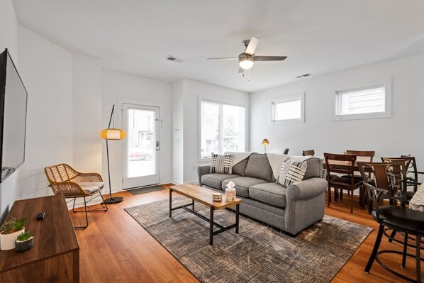 Open living room with a plush couch and a ton of natural light; containing a Roku flat-screen TV and electric adjustable desk.