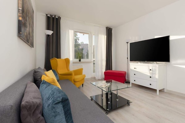 A bright living room with a grey sofa, a yellow armchair, a white sideboard, and a large flat-screen TV. Big windows let in plenty of natural light.