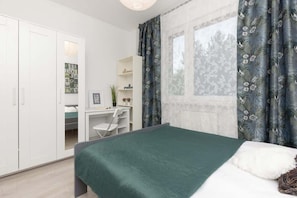 A bedroom with a green bedspread, white furniture, and a wardrobe. Tall windows offer an outdoor view.
