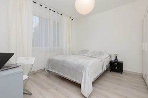 A cozy bedroom featuring a double bed with white linens, a bedside table, and sheer curtains allowing light to come through.