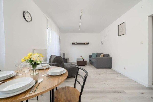 A modern kitchen and dining area featuring a dark fridge, a round dining table with a vase of flowers, and bright windows letting in natural light.

