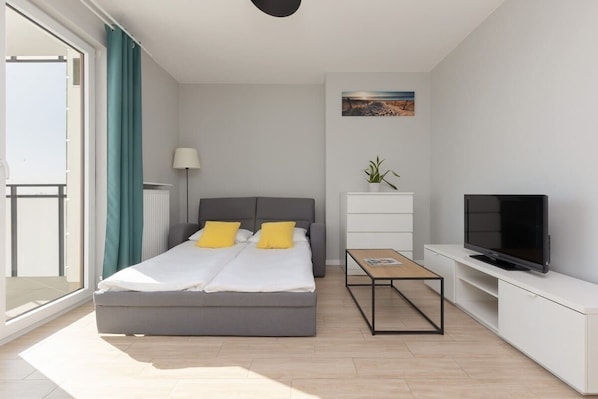 A bright bedroom featuring a large bed with yellow accents, a wall-mounted TV, and a modern, minimalistic design.