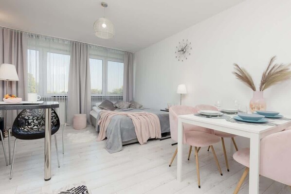 A bright living and dining area with large windows, light wooden flooring, a pastel pink dining set, and modern decor.
