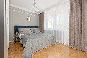 A bedroom with a large bed and soft gray bedding. The room is neatly decorated with light tones and minimal furniture, creating a serene atmosphere.