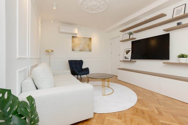 A bright living room with white walls, a light-colored sofa, and wooden floors. A small round coffee table sits in the center of the room, adding to its modern, airy feel.