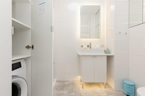 A clean bathroom with white tiles, a vanity unit, and a washing machine, offering both style and practicality.
