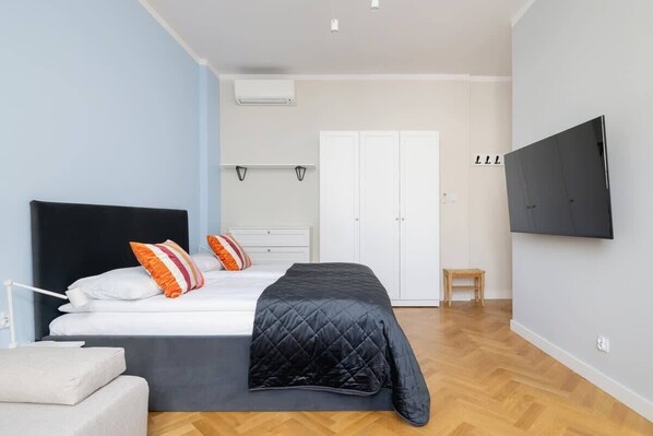 A cozy studio with a bed, sofa, wall-mounted TV, and pastel blue walls for a calming ambiance.
