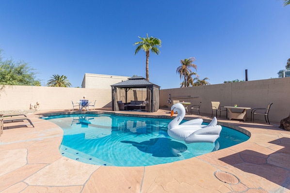 Spacious Entertainer's Backyard featuring a sparkling blue pool, hot tub, built-in BBQ, fire pit, lanai gazebo, putting green, half-court basketball, outdoor dining, and a large grass area perfect for yards games and furry friends.