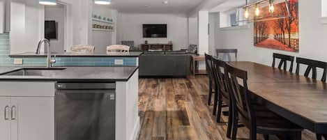 An open-concept kitchen and dining area with a sink and dishwasher on the left, a long dining table with chairs on the right, and a seating area with a sofa and TV in the background.