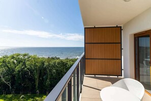 A balcony with a table and chairs, offering a relaxing spot with a scenic view of greenery and the coastline.
