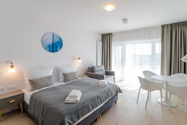 A bright bedroom with a double bed covered in gray bedding, bedside lamps, and a large window offering plenty of natural light.
