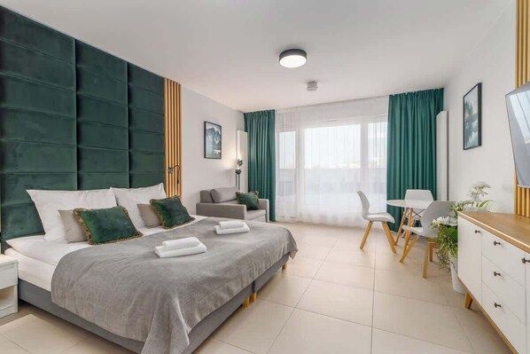 A spacious bedroom with a double bed, gray bedding, and green accents, complemented by natural light and modern decor.
