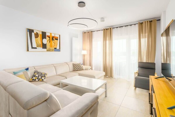 A bright living room with a comfortable sofa, a small coffee table, and large windows with sheer curtains, creating a cozy and airy space.
