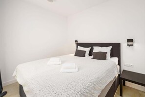 A minimalist bedroom featuring a double bed with white bedding, gray cushions, and bedside lighting, emphasizing simplicity and comfort.

