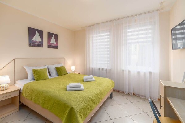 A cozy bedroom with a green bedspread, soft lighting, and sheer curtains allowing natural light.

