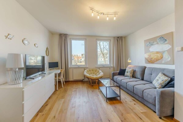 A spacious living room with a gray sofa, a white TV stand, and large windows allowing natural light to fill the space.
