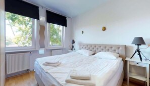 A cozy bedroom featuring a double bed, soft lighting, and a large window with dark curtains.
