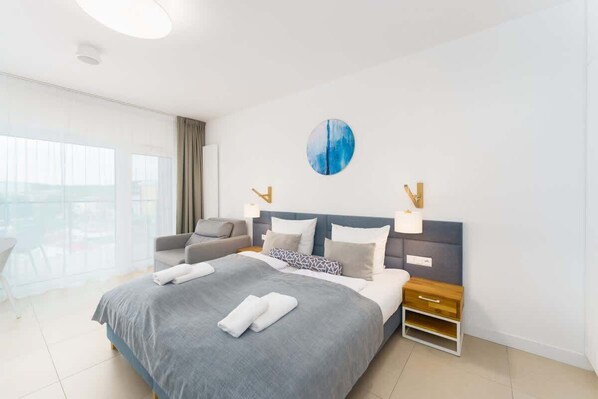 A bright bedroom with duble bed, blue accents, and floor-to-ceiling windows leading to a balcony.
