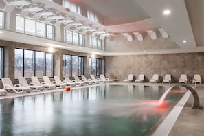 An indoor swimming pool with lounge chairs and large windows offering ample natural light.
