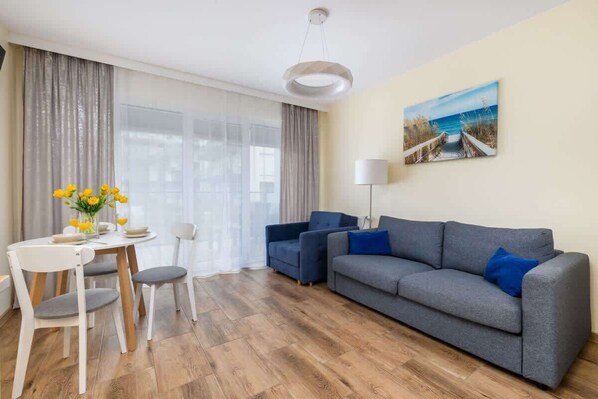 A bright living room with a blue sofa, a white coffee table, and a dining area near large windows.
