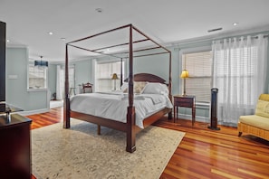 A serene bedroom space with a dominant four-poster king bed. The hardwood flooring complements the wooden furniture in the room. There's a gentle touch of colour with the pale blue walls and the white bedding. Bedside tables with lamps flank the bed, providing additional light sources. The room has multiple windows, ensuring ample natural light.