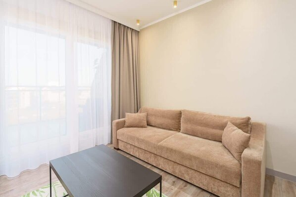 A cozy living room with a beige sofa, a glass coffee table, and natural light streaming in through large windows.
