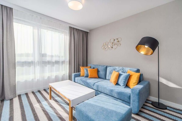 A modern living area featuring a blue sofa with orange and white cushions, a striped rug, and natural light from large windows.

