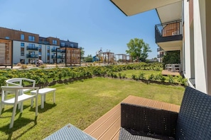 A sunny ground-floor terrace featuring a grassy area, outdoor seating, and neighboring apartment buildings in view.

