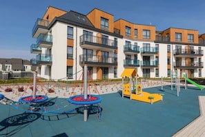A contemporary apartment complex near the waterfront, complete with balconies and a colorful playground.


