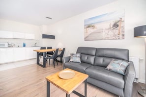 A modern open-plan living area with a gray sofa, wooden coffee table, and a seaside painting, set on light wood flooring.

