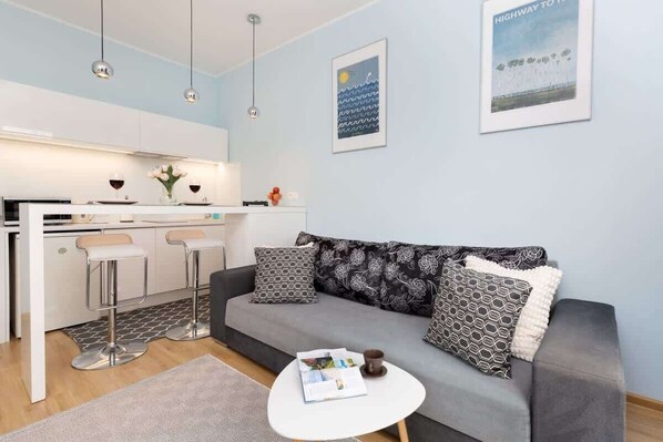 A cozy living area with a grey sofa, a small round coffee table, and a kitchenette in the background.
