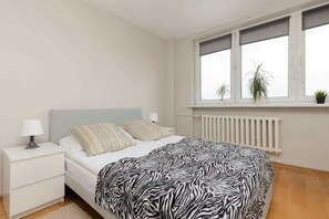 A minimalist bedroom with a white bed, a zebra-patterned throw, and natural light from the windows.
