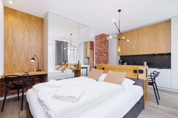 A wide perspective of a modern apartment with elements of brick, wood and black. In the centre stands a double bed behind which you can see the kitchenette. 