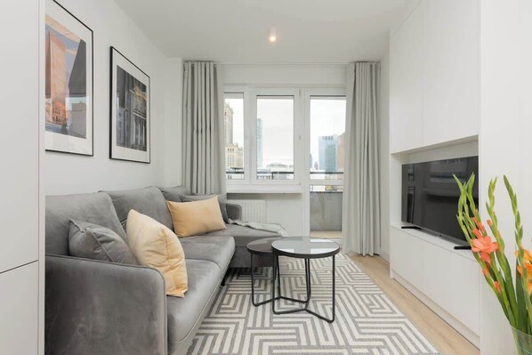A bright living room with a gray sofa, yellow accent pillows, a modern TV, and a small balcony.
