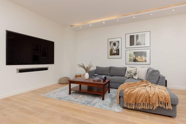 A cozy living room with a modern gray sofa, a coffee table, and a large TV on the wall.
