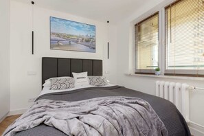 A cozy bedroom with a double bed, gray bedding, a cityscape artwork, and a large window with blinds.
