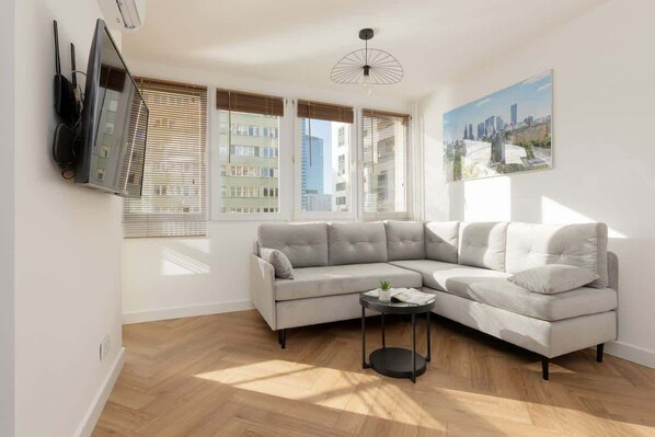 A modern living room with a gray sectional sofa, a TV mounted on the wall, and large windows letting in natural light.
